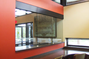A new common space at Rockdale Temple. Chairs are tucked into counter along the wall of a work space. One wall features a glass section, allowing those in the work space to see into the foyer. Just past the glass section can be seen a stained glass window featuring Hebrew script is on display in a case and hanging on the wall.