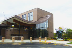 The exterior of the main entrance to Rockdale Temple. The building is brown brick. The roof of the foyer is a eye-catching U shape, sloping down to the door, then curving back up as it extends out to cover the outdoor entry way. The foyer is all glass. Large cement bollards line the curb, painted beige. To the right is a large sculpture of figures playing instruments surrounded by a small garden of shrubs. Trees and plants are in the background.