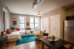 A cozy studio apartment. Two twin beds are tucked in the corner with white bed spreads and vibrant colored and patterned throw pillows. A pale yellow arm chair is in the center of the room facing the beds. Two small round tables with metal supports and glass tops accompany the furniture. There are two large windows on the far wall, allowing lots of natural light. A small table with seating for two can be seen in the bottom right corner, with a decorative plant in the center. Closet doors line the far right wall. The walls are taupe. An exposed duct is on the ceiling. To the far right is a stainless steel refrigerator. A bright teal rug is in the center of the room.