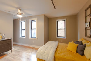 Bedroom of Republic Lofts Apartments. Queen bed with bright yellow bedding hugs the right wall. Hardwood floors and three windows letting in natural light. A ceiling fan and light hangs from the ceiling at the left side of the room. A small dresser is against the left wall. The walls are painted a neutral taupe.