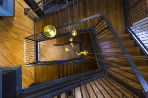 A view looking down a three-story wooden staircase with steel railings. The staircase makes a rectangular shape as it twists downward. The wood is plain-sawn white oak, laminated. Lights dangle through the center of the stairs, appearing as sparkling golden-yellow orbs.