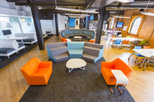 A light-filled and bright and colorful modern office space, adapted in an old warehouse. The wall to the right is exposed bright with arched windows surrounded by a two-toned wooden frame. Large rectangular tables are surrounded by eight chairs colored sky blue, vivid orange, medium gray, and lime green. The left wall is plastic with white designs printed. This wall has booth seating with benches covered in graphic patterned upholstery. The center area has a wide strip of gray carpet over hard wood floors. The carpeted area has two orange arm chairs in the foreground, two gray and blue staircase-style chairs, then two more orange arm chairs facing a large circular upholstered bench. The office space extends past this common area, and there is even a kitchen in the far right corner.
