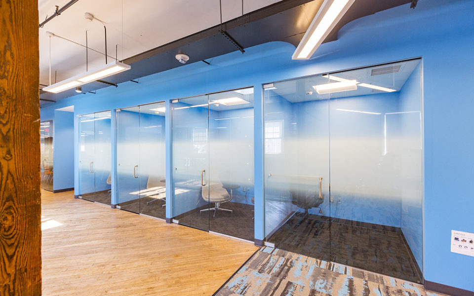 A blue wall with four glass doors leading into private phone call spaces as part of a modern office space. The rooms have dark gray carpet, and each room has a chair and an outlet. The right side of the hallway has a brightly pattern carpet, which ends and begins a hard wood floor hallway. A wood beam frames the left side of the image. Fluorescent lighting hangs from a high, white ceiling.