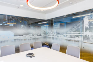 A conference room. The room is enclosed by glass walls, but the glass has the Cincinnati skyline imprinted on it in a white-scale color scheme. The conference table is large and square, with cool gray chairs pushed in. A circular light with orange accent casing hangs from the white ceiling. A large staircase can be seen beyond the conference room walls.