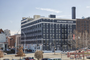 The Strietmann Center. A large, historic building at the corner of a mid-sized city intersection. The building is brick, painted a dark blue-gray. There are six stories, each either lots of windows. The windows have white stone above it. The sky is blue with one wisp of a cloud above the building.