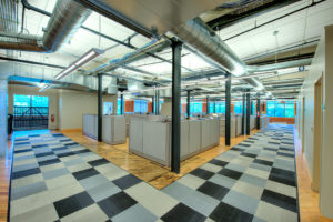 An open office space. A cluster of cubicles are centered in a large open space with hardwood floors. Gray checkered rugs mark the walkway around the cubicle space. The back walls have large windows, looking out on green wooded space. The exterior walls are exposed red brick and the window frames are rustic wood. The ceiling displays exposed ducts. Support beams throughout the room are black. Fluorescent lighting illuminates the bright space.