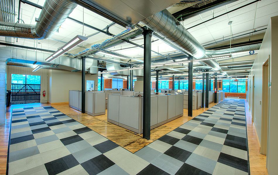 An open office space. A cluster of cubicles are centered in a large open space with hardwood floors. Gray checkered rugs mark the walkway around the cubicle space. The back walls have large windows, looking out on green wooded space. The exterior walls are exposed red brick and the window frames are rustic wood. The ceiling displays exposed ducts. Support beams throughout the room are black. Fluorescent lighting illuminates the bright space.
