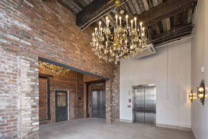 The lobby of a renovated building. Exposed brick walls and aged wooden ceilings show the building's history, but two brad-new elevators are at the far wall, and two grand chandeliers hang from the ceilings.