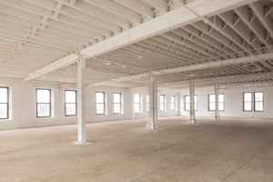 A renovated but unfinished space in a large historic building, a former factory. The far wall and left wall are lined with large windows. Steel columns cross the middle of the room. Ceiling beams are exposed, but painted a clean white.