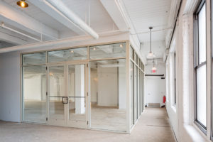 Potential office space in a newly renovated building, originally an historic factory. Painted all white, a hallway runs along the window-filled exterior wall to the right. To the left is a room, framed by glass walls to let in the natural light. Double doors face the camera, as the hallway continues left off camera.