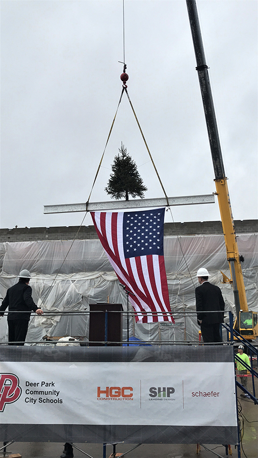 A large yellow crane lifts a ceremonial beam onto the top of Amity Elementary. The beam also holds a small fir tree, and an American flag hangs from the bottom of the beam.