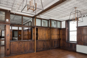 A potential conference room with dark wood paneling, renovated from an historic factory. The far wall features built-in cabinetry, and the top row of paneling has been replaced with glass to let light pour through the building. The ceiling is painted white and features crown molding. The floors are a dark wood to match the panels on the walls.