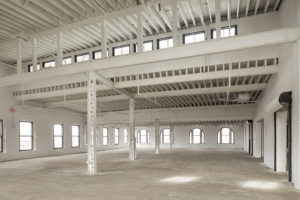 A renovated but unfinished space in a large historic building, a former factory. Large doorways with overhead doors are along the right wall. The far wall and left wall are lined with large windows. Steel columns cross the middle of the room. Ceiling beams are exposed, but painted a clean white.