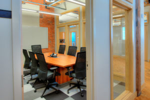 A small conference room. The walls are windowed, making the space feel bigger and brighter. A wooden conference table with one phone in the center takes up most of the room. Eight large black office chairs surround the table. A dry erase board is on the far wall, which shows some exposed red brick. Gray checkered rug covers the floor. The windows are framed in light wood, and the walls are a very pale gray, almost white.