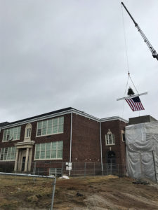 A large crane lifts a ceremonial beam onto the top of Amity Elementary. The beam also holds a small fir tree, and an American flag hangs from the bottom of the beam. The front of the building is brick with nine-pane windows, and a double-door entrance bedecked by stone columns.