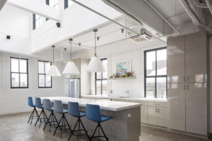 A very clean and chic modern kitchen. The walls, ceilings, counters, lighting, and cabinets are all very white. Lots of windows let in cool light. Six tall blue chairs are at the bar. A shelf over the sing holds rustic knick knacks and a colorful painting.
