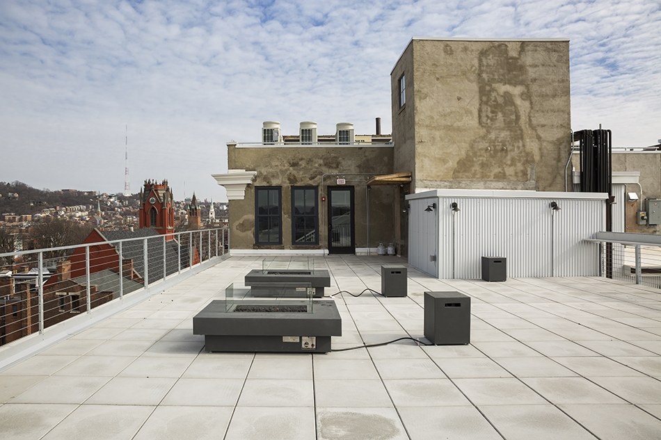 The rooftop deck of a building, with two square electric fire pits surrounded by bench seating. The entrance to the roof top is in the background, a cement structure with new, tall windows and a glass door. A view of a city beyond the railing to the left. Blue skies peek through a cottony layer of clouds.