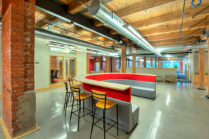 An open office space, mixing the aesthetics of modern and rustic. A large U-shaped piece of furniture is in the center. The inside is upholstered in gray and red and serves as a bench, the top has a shelf extending around it, allowing the piece of furniture to double as a bar. Tall goldenrod stools sit around the bar. In the distance are beige arm chairs, wooden support beans, and lots of windows.