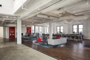 A large, modern, corporate gathering space. The far left wall holds two bright red book shelves, and a sitting area. A second sitting area is closer to the camera. An extremely long conference table lines the far right wall, with rolling desk chairs tucked in. Most of the floor is hardwood, except for a section to the left which is cement. There are lots of windows, and almost everything is white, including walls, ceilings,exposed duct work, and columns.