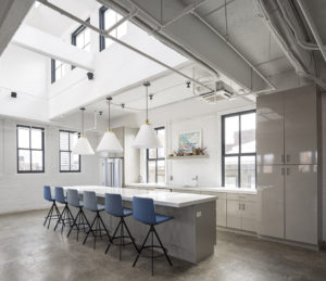 A very clean and chic modern kitchen. The walls, ceilings, counters, lighting, and cabinets are all very white. Lots of windows let in cool light. Six tall blue chairs are at the bar. A shelf over the sing holds rustic knick knacks and a colorful painting.