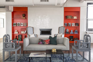 A vibrant, modern sitting area. The far wall holds a small fireplace tucked into a white painted brick wall, with bright red floor-to-ceiling bookshelves on either side. Two high-backed arm chairs in a soft sky blue sit to either side of the fireplace. A long, gray sofa faces the camera. Two throw pillows sit in the middle of the couch. Two ornately carved chairs, which were painted gray in this renovation, are on either side of the sofa. Two coffee tables are in the center, with a bellhop bell and some pamphlets. A decoratively patterned blue rug is under the couch and carved chairs.