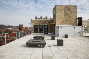 The rooftop deck of a building, with two square electric fire pits surrounded by bench seating. The entrance to the roof top is in the background, a cement structure with new, tall windows and a glass door. A view of a city beyond the railing to the left. Blue skies peek through a cottony layer of clouds.
