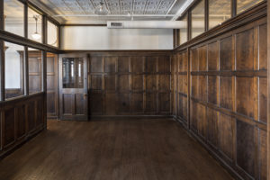 A potential conference room with dark wood paneling. The left wall has windows looking into the hall and letting in light. The door has a large window, and is propped ajar. The ceiling is painted white and features crown molding. The ducts are exposed, but painted white to blend in with the ceiling. The floors are a dark wood to match the panels on the walls.