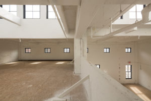 Interior area of a recently renovated historic building. Formerly a factory, the space is now ready for occupancy. This two-level loft space features high ceilings with windows near the ceiling. An upper floor can be seen to the left, with a half wall leaving the space open to the lower floor to the right. The space is white, the floors are bare plywood for now. Pipes and duct work are exposed at the ceiling, but painted white.