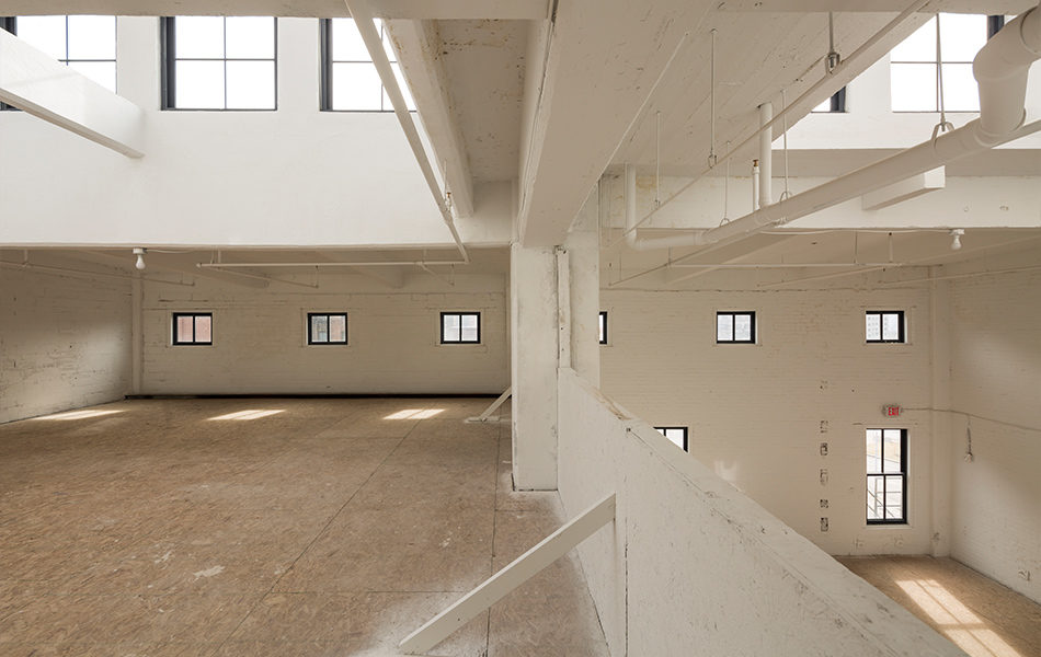 Interior area of a recently renovated historic building. Formerly a factory, the space is now ready for occupancy. This two-level loft space features high ceilings with windows near the ceiling. An upper floor can be seen to the left, with a half wall leaving the space open to the lower floor to the right. The space is white, the floors are bare plywood for now. Pipes and duct work are exposed at the ceiling, but painted white.