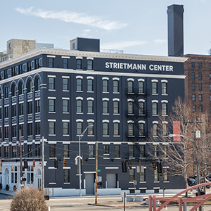 The Strietmann Center. A large, historic building at the corner of a mid-sized city intersection. The building is brick, painted a dark blue-gray, except for the street-level. At the street level, the side with lots of windows and storefront entrances is painted white, while the side facing the camera and having smaller windows and only one entrance is the blue-gray painted brick. There are six stories, each with lots of windows. The windows have white stone above it. The sky is blue with one wisp of a cloud above the building.