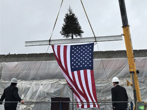 A large yellow crane lifts a ceremonial beam onto the top of Amity Elementary. The beam also holds a small fir tree, and an American flag hangs from the bottom of the beam. Two men in dark suits and light hard hats watch, their backs to the camera.