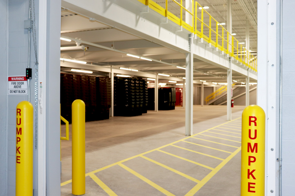 The doorway into a warehouse that stores dumpsters. The doorway is framed by yellow bollards with "Rumpke" printed on them in red. Through the doorway is a two-story warehouse. The ground level has stacks and stacks of dumpsters. The second story has a yellow railing to allow viewing to the first floor. A yellow-striped pedestrian walkway leads from the doorway through the warehouse.