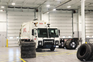 A large white trash truck sitting in a warehouse garage. The garage door is closed behind it. A yellow painted pedestrian walkway crosses in front of the truck. Four tires are stacked next to the walkway, and more tires lean on racks to the right of the truck.