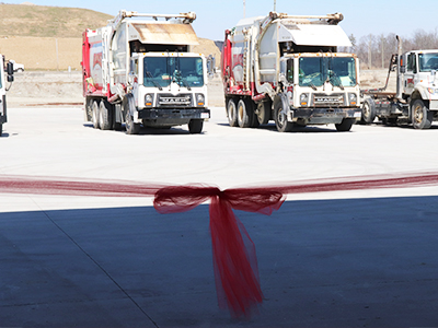 A red ribbon is tied in a bow, and is strung across the center of the image frame. In the distance are two white garbage trucks and one white Mack truck with an empty truck bed. The foreground is in the shadows, while the trucks sit in the sunlight. The sky is blue, and a brown hill can be seen in the distance.