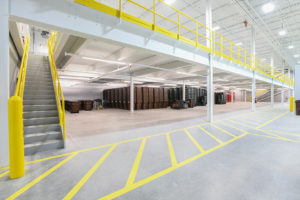A large industrial garage for storing trash and recycling bins. The entire facility is very gray, with cement floors and corrugated metal roofing. Fluorescent lighting. Yellow stripes indicate the pedestrian walkways within the facility, and in the far left is a staircase leading to second floor. The stair railing is bright yellow, as is the second floor railing, which can be seen from the first floor.