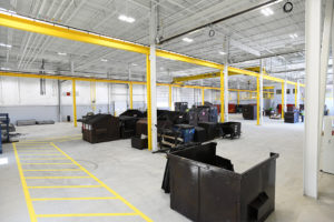 A large industrial garage for repairing trash and recycling trucks. The entire facility is very gray, with cement floors and corrugated metal roofing. Fluorescent lighting. A pedestrian walkway, painted yellow on the floor, heads to the left of the frame and then turns to the right. Large brown dumpsters are placed throughout the warehouse space, to be cleaned or repaired.