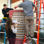 Three people are assembling a cylindrical structure out of canned goods.