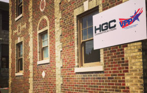 A building, with mainly red brick and some gray brick creating decorative patterns. Windows, framed by gray brick. A sign hangs on the wall in the top right corner of the picture, displaying the HGC Construction logo and the Voluntary Protection Programs logo.