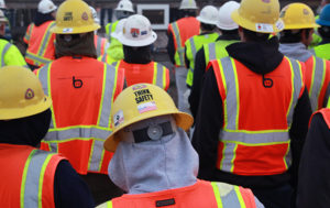 A group of construction workers look away from the camera, wearing hard hats, sweatshirts, and orange hi-visibility safety vests. The person centered in the foreground has a stick on their hat that says, "THINK SAFETY."