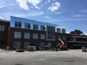 A modern three-story red brick school building, Amity Elementary, under construction with large equipment in front. A sunny day with blue sky and wispy clouds.