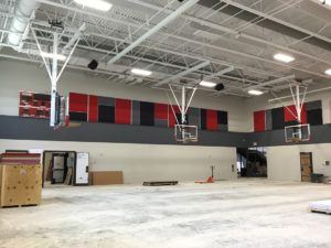 A school gymnasium under construction at Amity Elementary. The floor is unfinished and dusty, but the basketball hoops are installed and red gray and black wall padding adorns the walls.