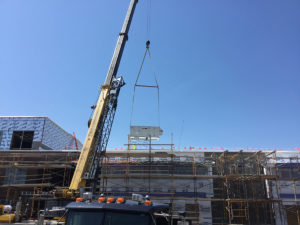 A large yellow crane lifts a hefty HVAC machine onto the roof of the new addition for Amity Elementary. The front of the building is covered in construction scaffolding, and the top of a truck can be seen in the nearest foreground. The sky is blue with not a cloud to be seen.