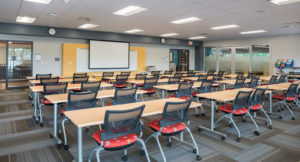 A large classroom, seen from the back of the room. Four rows of tables span the classroom, and each table has two rolling chairs with bright red patterned upholstered cushions tucked beneath the table. The front of the classroom has an accent of yellow, and a whiteboard on display. This room is for teachers professional development.