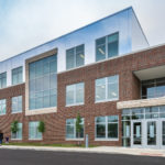 The new entrance to Amity Elementary. The three-story structure has clean lines and large windows. The first two stories have a facade of red brick, and the third story has a facade of mirrors so that the top story seems to connect with the sky. There are ribbons of beige stone accents.