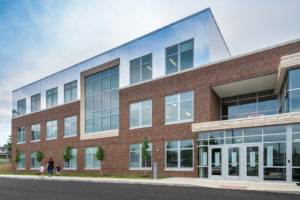 The new entrance to Amity Elementary. The three-story structure has clean lines and large windows. The first two stories have a facade of red brick, and the third story has a facade of mirrors so that the top story seems to connect with the sky. There are ribbons of beige stone accents.