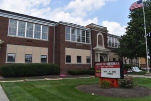 The front of Amity Elementary prior to the renovations of Summer 2018. A red sign surrounded by a circular garden on the front lawn, boasts of "Deer Park Pride" and tells students to "Have a safe summer." The first floor of windows feature small, single-pane windows surrounded by beige fill in, in contrast to the second floor window which have more traditional looking, 9-panel windows. An arch of decorative stone is above the arched front entrance, which is beneath a small stone overhang supported by stone columns. The front of the overhang has "Amity Elementary School" displayed in black iron lettering. An American flag waves in the wind on the right side of the image. The sky is blue and there are fluffy white clouds.