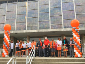 A group of high school girls and middle-aged to elderly adults stand in front of a large building, as part of the ceremonial Mercy McAuley ribbon cutting. There are columns of orange gray and white balloons. 