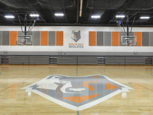 A school gymnasium. The wolf mascot is painted on the center of the court in gray, white and orange. The mascot image is also painted on the wall, with "Mercy McAuley Wolves" printed beneath, and surrounded by gray and orange wall padding. There are two basketball hoops, one on either side of the frame. The gray bleachers are in the background, tucked away. The ceiling is black.