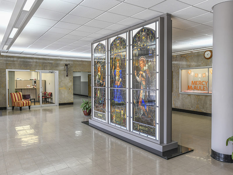 An historic three-paneled stained glass window on display in a school lobby. The stained glass depicts the Mother Mary in the center, holding the baby Jesus, with an angel on either side. The school lobby is well-lit, with an overall cool gray coloring, and a chair outside of the office entrance. The chair is upholstered in red and yellow striped fabric. There are two potted plants in the lobby.