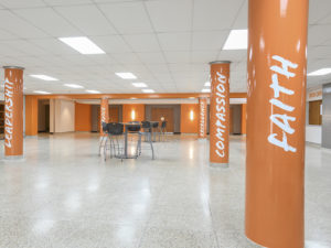 The lobby outside a school auditorium. Five columns are painted bright orange, and have the school values painted on them. From left to right, the columns read, "Leadership," "Service," "Excellence," "Compassion," "Faith." There are two bistro tables in the middle with four black chairs at each of them. The box office is to the far right. The floor is a cool gray, the walls a cheery orange, and a few accent walls are gray or white. Fluorescent lighting is on the ceiling.